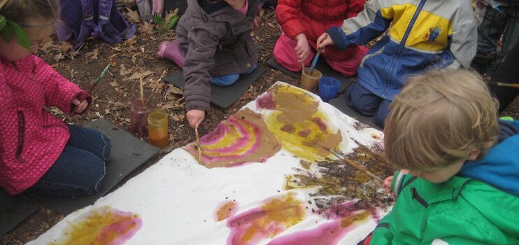 Eine Kindergruppe sitzt auf dem Waldboden und malt auf einer ausgebreiteten Stoffbahn