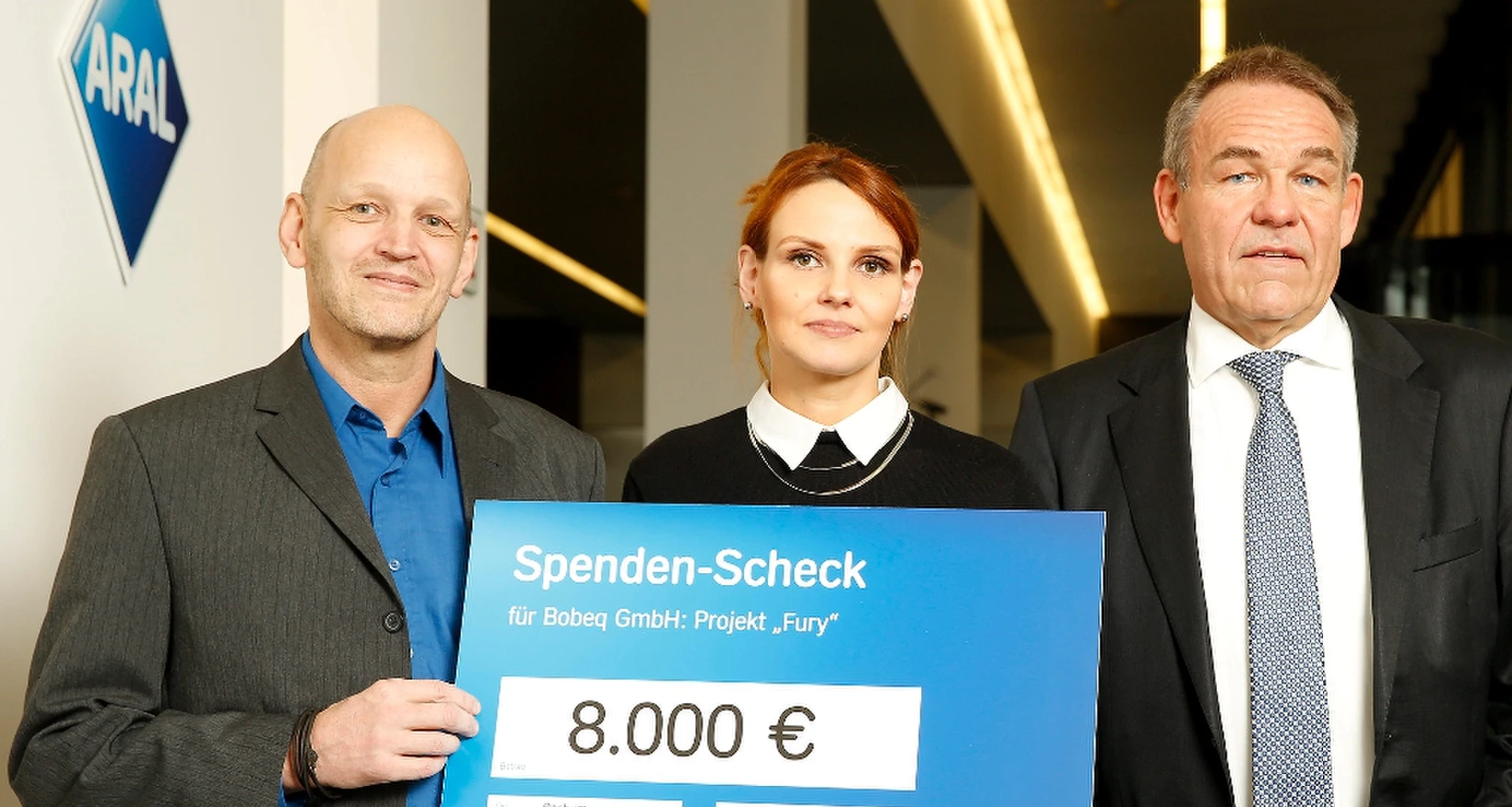 Zwei Männer und eine Frau halten einen symbolischen Scheck vor sich.