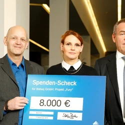 Zwei Männer und eine Frau halten einen symbolischen Scheck vor sich.