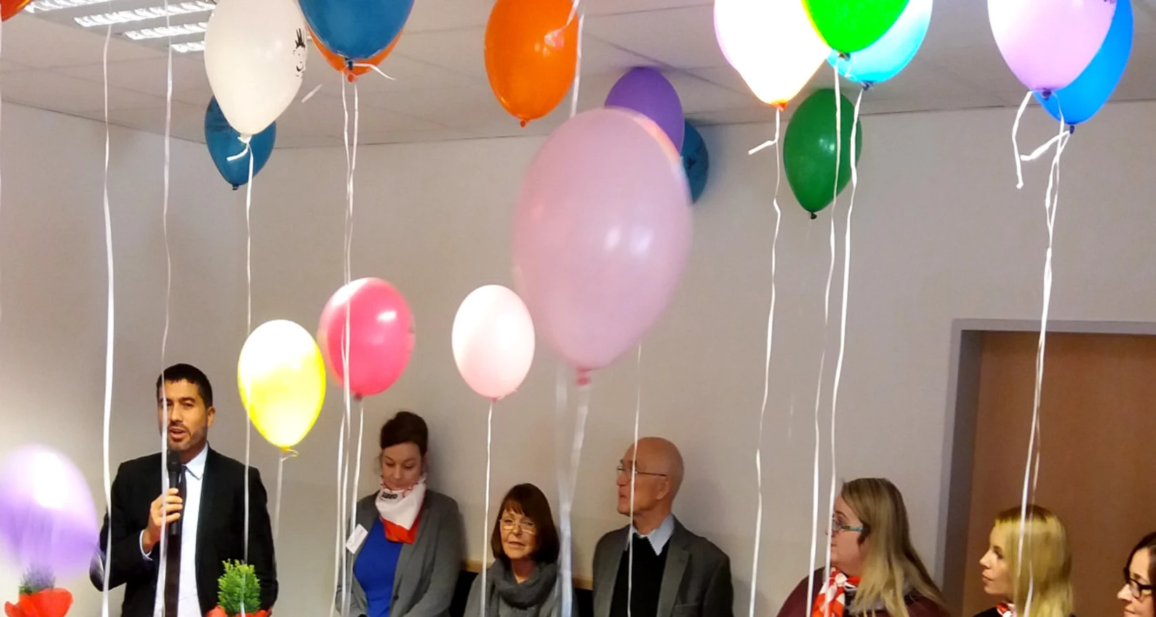Grußworte des Vorsitzenden an die Gäste der Kita-Eröffnungsfeier. Im Vordergrund Kinder, dahinter erwachsene Gäste. Luftballons schweben unter der Decke.