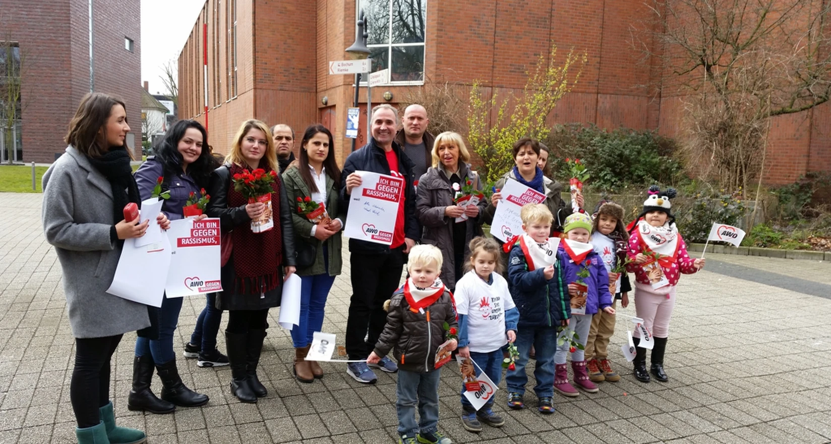 Menschengruppe (Mann, Frauen und Kinder) mit Plakaten "Gegen rassismus" und Rosen in der Hand vor einem Gebäude.