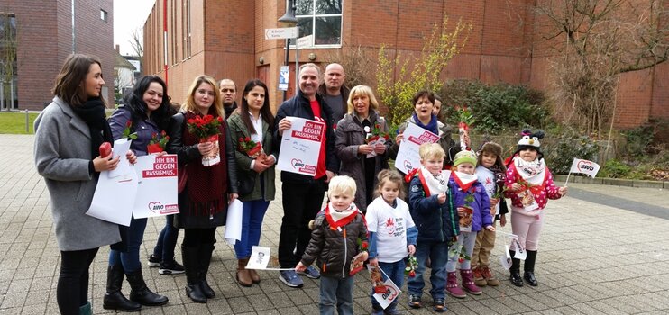 Menschengruppe (Mann, Frauen und Kinder) mit Plakaten "Gegen rassismus" und Rosen in der Hand vor einem Gebäude.