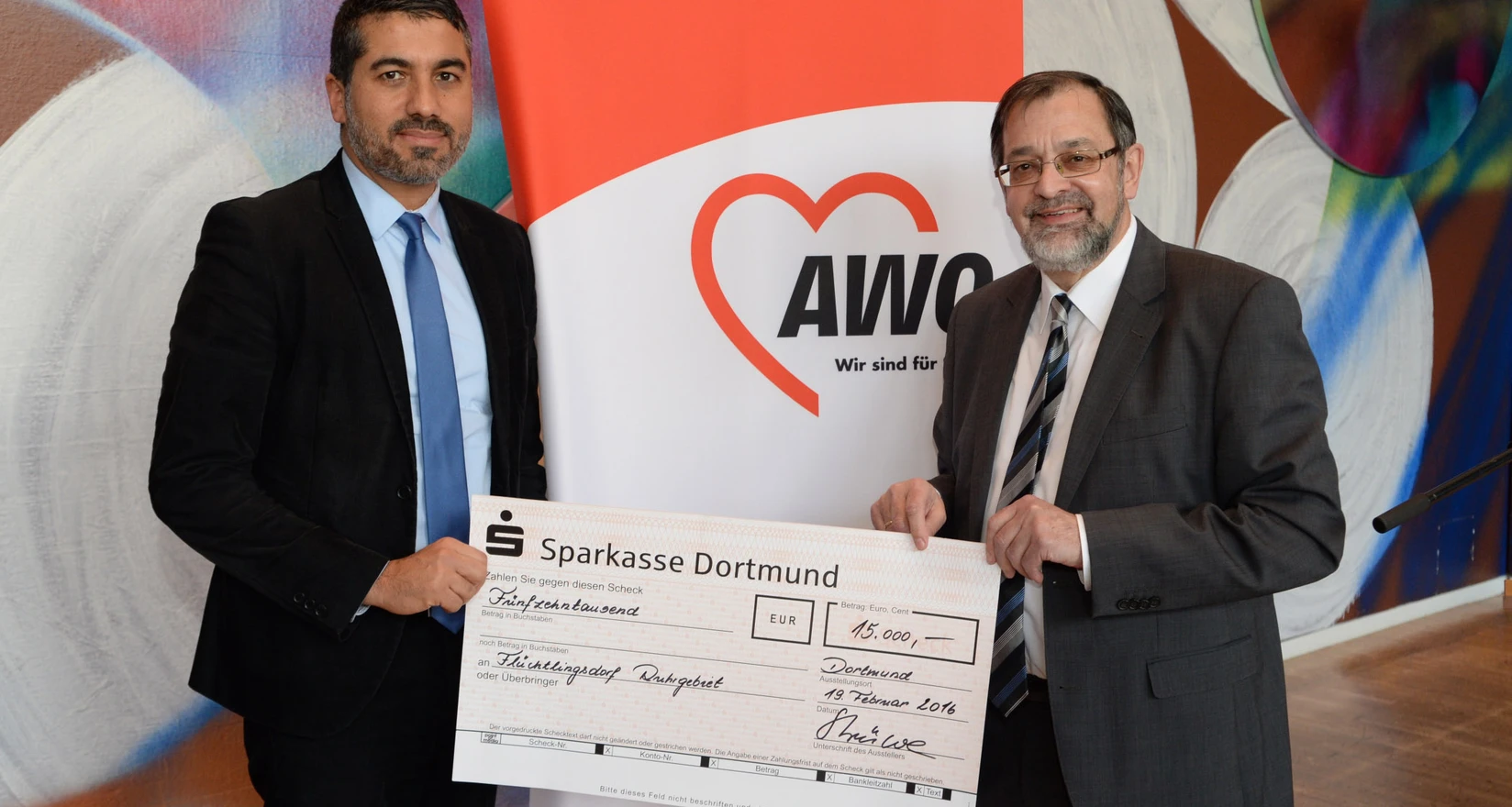 Links im Bild Serdar Yüksel (Vorsitzender UB Ruhr-Mitte, rechts im Bild Michael Scheffler (Vorsitzender AWO Bezirk Westl. Westfalen), beide halten einen symbolischen Scheck in der Hand. Im Hintergrund das AWO-Logo.