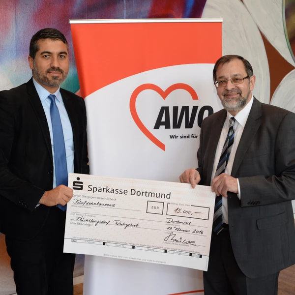 Links im Bild Serdar Yüksel (Vorsitzender UB Ruhr-Mitte, rechts im Bild Michael Scheffler (Vorsitzender AWO Bezirk Westl. Westfalen), beide halten einen symbolischen Scheck in der Hand. Im Hintergrund das AWO-Logo.