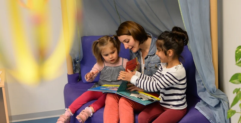 Zwei Mädchen mit Erzieherin schauen ein Buch an
