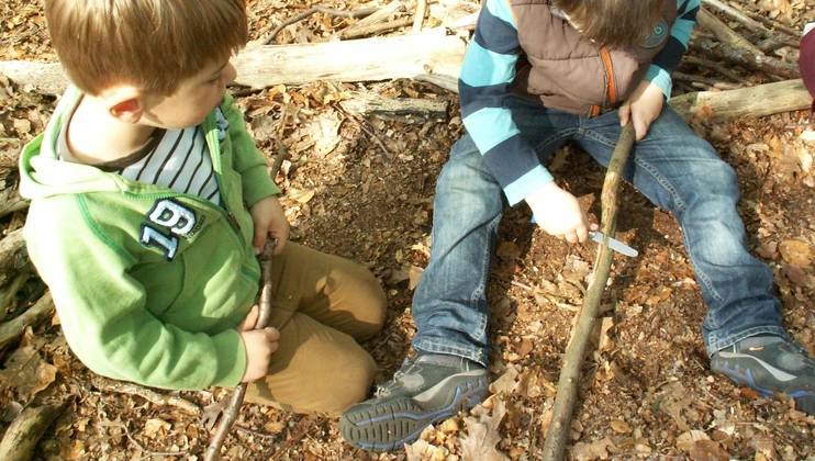 Zwei Jungen sitzen auf dem Waldboden, einer schnitzt an einem Ast