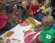 Eine Kindergruppe sitzt auf dem Waldboden und malt auf einer ausgebreiteten Stoffbahn