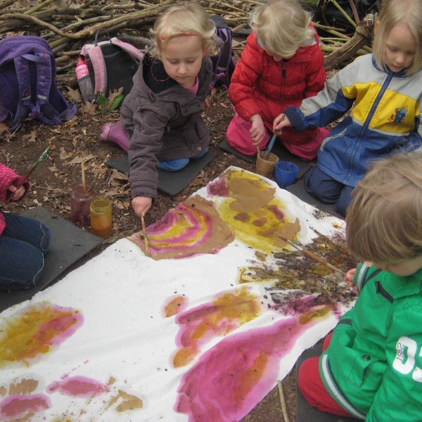 Eine Kindergruppe sitzt auf dem Waldboden und malt auf einer ausgebreiteten Stoffbahn