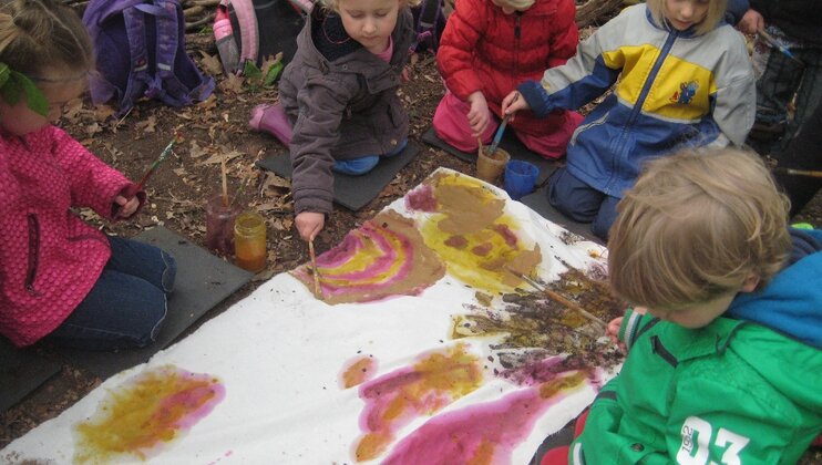 Eine Kindergruppe sitzt auf dem Waldboden und malt auf einer ausgebreiteten Stoffbahn
