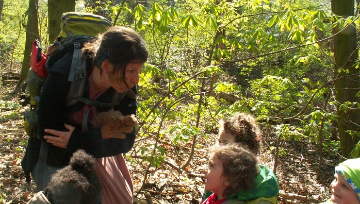 Eine Frau und Kinder im Wald - die Frau macht ewas vor und die Kinder hören lachend zu