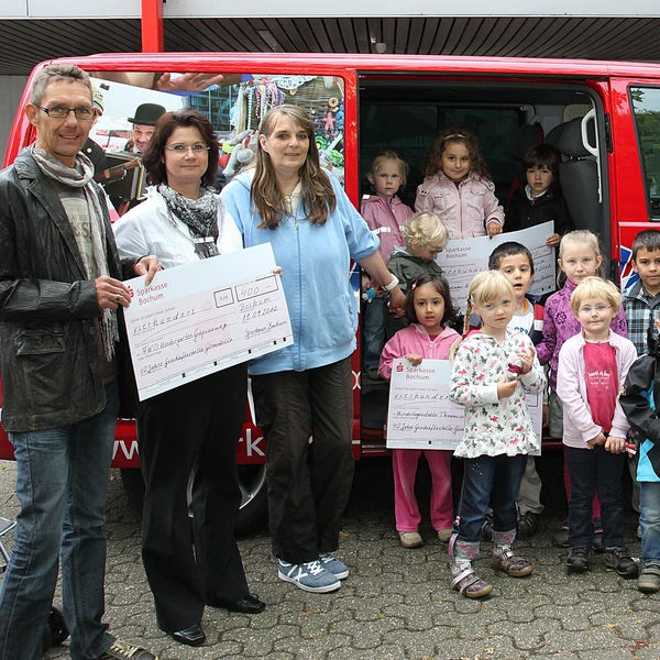 Kinder und Erwachsene mit symbolischen Schecks vor einem Kleinbus