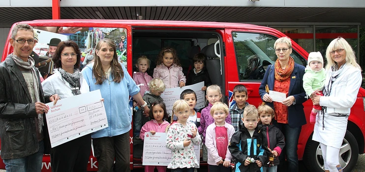 Kinder und Erwachsene mit symbolischen Schecks vor einem Kleinbus