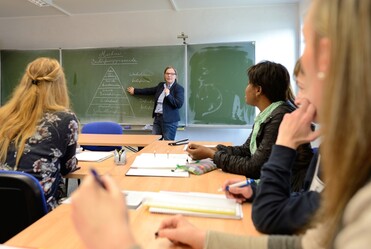 Unterricht: eine Frau zeigt den vor ihr sitzenden Schülerinnen ein Schaubild an der Tafel