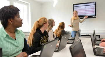 Schülerinnen sitzen vor ihren Laptobs und schauen auf eine Frau, die ewas an winwm Wandbildschirm erklärt.