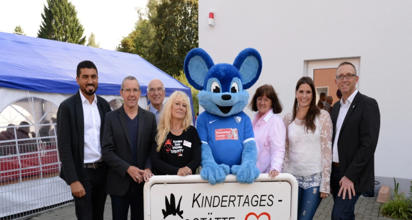 Ehrengäste, Geschäftsführung, Pädagogische Leitung und Kitaleitung vor dem Eingangsschild der Kita. In der Mitte eine blaue Riesenmaus.