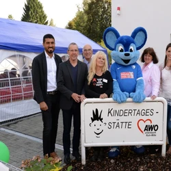 Ehrengäste, Geschäftsführung, Pädagogische Leitung und Kitaleitung vor dem Eingangsschild der Kita. In der Mitte eine blaue Riesenmaus.