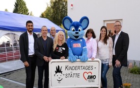 Ehrengäste, Geschäftsführung, Pädagogische Leitung und Kitaleitung vor dem Eingangsschild der Kita. In der Mitte eine blaue Riesenmaus.