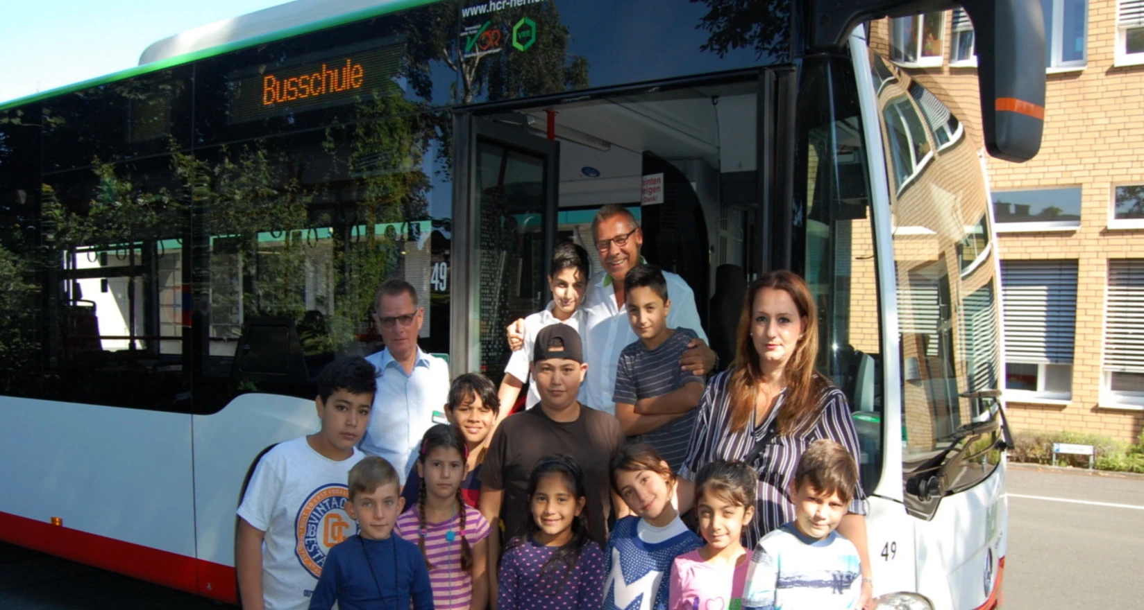 Menschengruppe (Erwachsenen und Kinder) vor einem Bus mit der Aufschrift Busschule