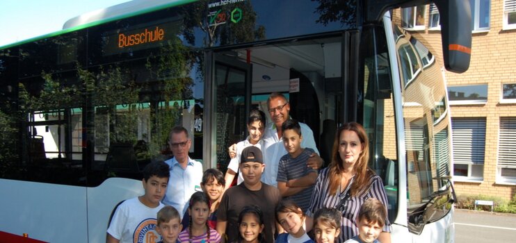 Menschengruppe (Erwachsenen und Kinder) vor einem Bus mit der Aufschrift Busschule