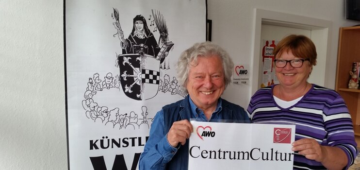 Karikaturist Waldemar Mandzel und Gabriela Osterkamp vor einem Banner des Künstlertreff WAT. Sie halten ein Bild mit der Aufschrift CentrumCultur in die Kamera.